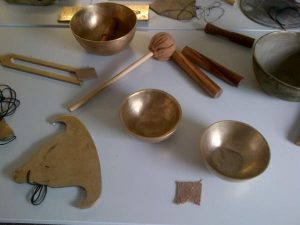 This image shows a table display of bronze bowls, wooden sticks, a cloth covered gong stick, and a flat bronze gong in a shape that resembles a bell, a Tibetan Lama's hat, or a Buddha.