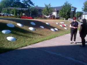 This image shows David Johnson's art installation 'Too Big To Feel' - 16 large white concrete mounds, and one red mound, set in the grassy embankment by a path on the Royal Holloway campus, to read, in Grade 2 contracted Braille, 'Seeing Red'. The artwork is seen here with two female conference delegates, one of whom has short, bright red hair