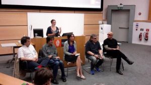 This image shows writers Frédéric Grellier, Romain Villet, Naomi Foyle, Rod Michalko and Ryan Knighton sitting on chairs arranged in a row in front of a long computer podium. Hannah Thompson is standing behind the podium, addressing the auditorium audience