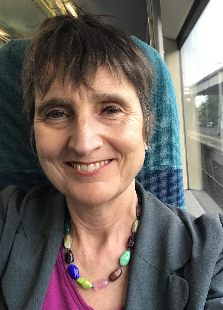 Naomi Foyle, a white woman with short brown hair, sitting in a train, wearing a necklace of colourful stones and smiling.
