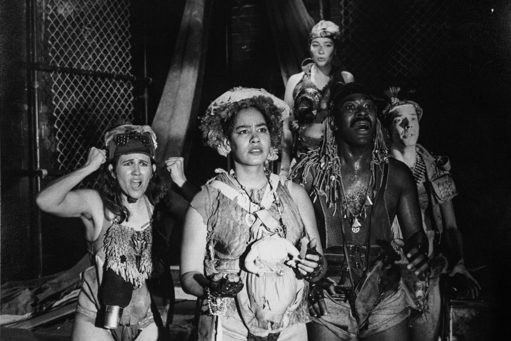 The cast of HUSH: a diverse group of performers dressed in rags, centred around a Black woman with a headless white baby doll strapped to her front. From L to R: Randi Helmers, Tabby Johnson, Holly Cole, Jimmie Earl Perry, James Tait.