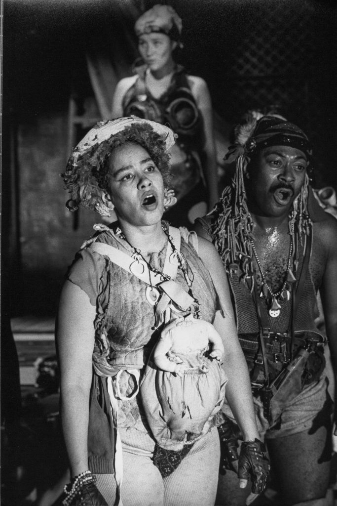Tabby Johnson and Jimmie Earl Perry singing, with Holly Cole as the Pied Piper of Hamelin, standing behind them. Tabby Johnson, a Black woman, has a headless white baby doll strapped to her front.