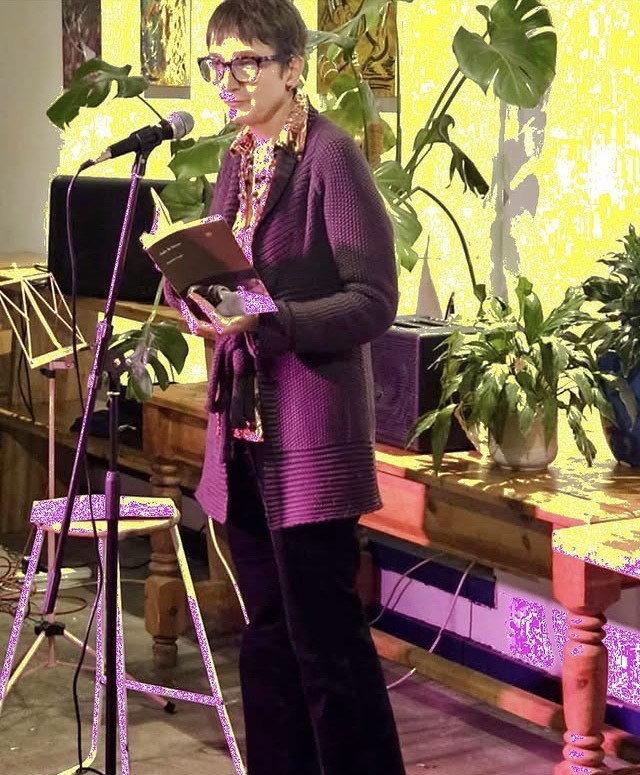 A photo of Naomi Foyle reading at a microphone with plants throwing shadows on the wall behind her. Digitally altered in technicolour to give a golden glow.